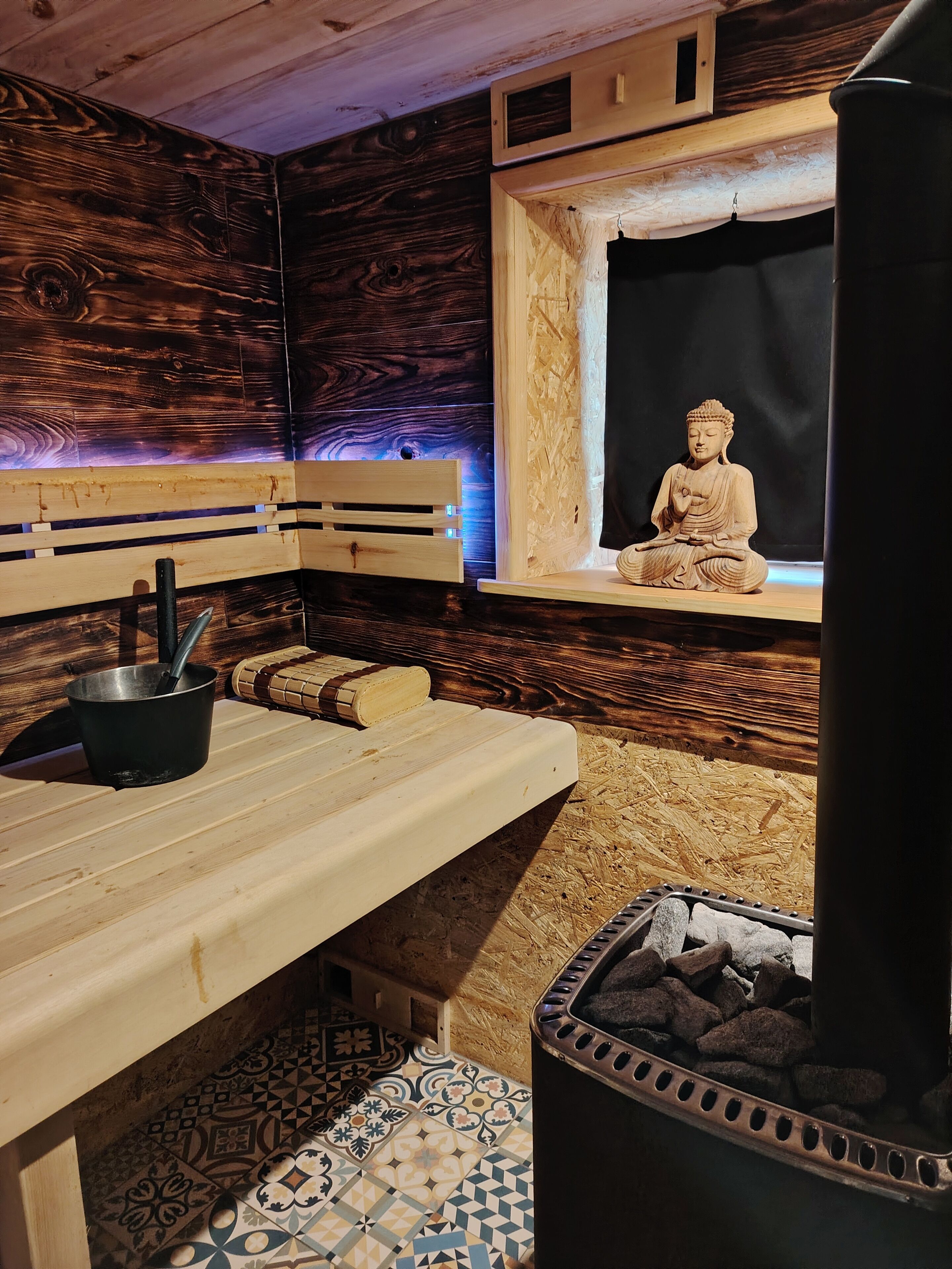 A sauna room with a wooden bench, a stone Buddha statue, a black pot, and a stone heater.