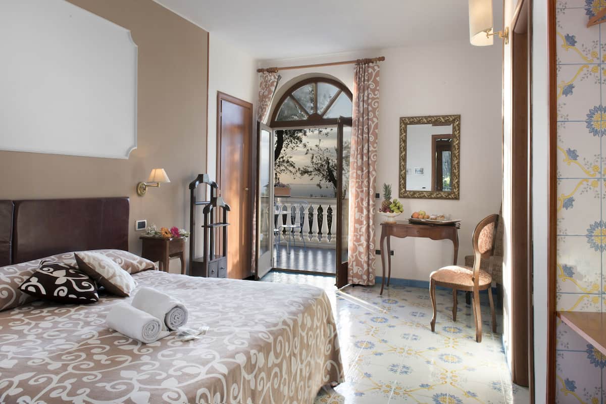 A hotel room with a bed, a desk, a chair, a mirror, and a view of the outside.