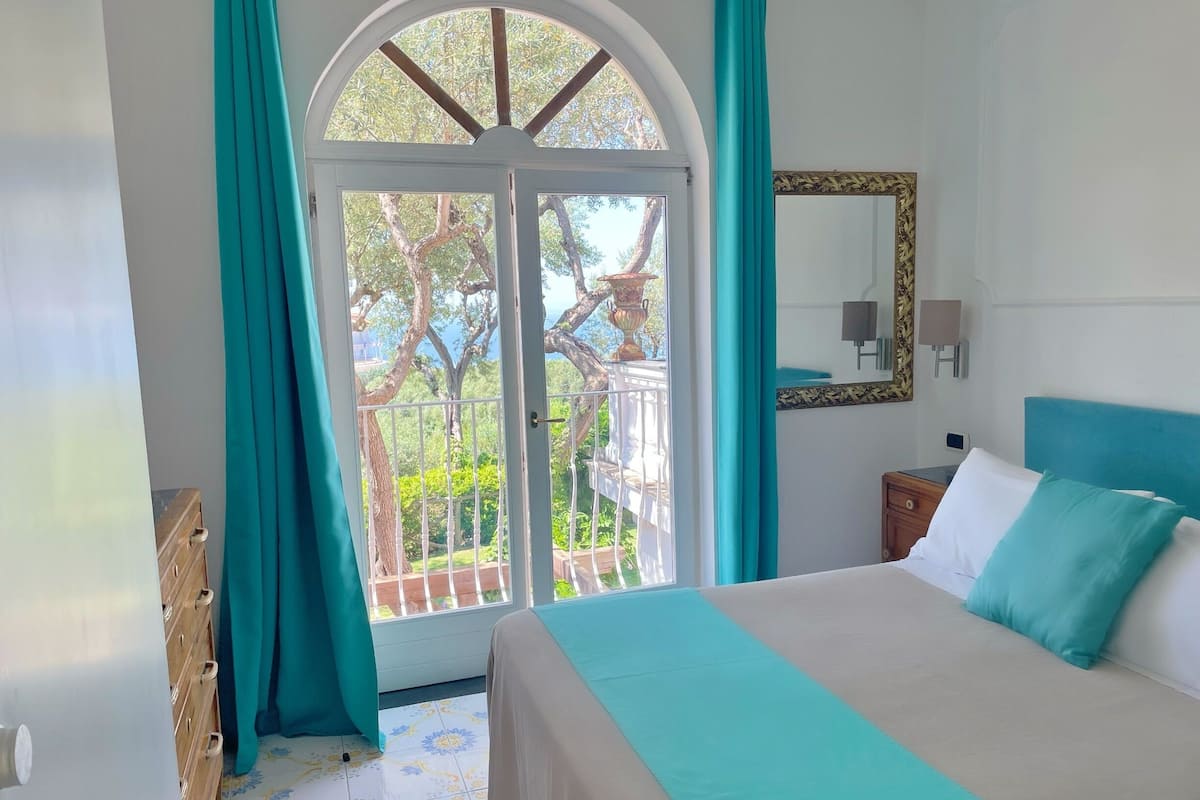 A bedroom with a bed, a dresser, a nightstand, a mirror, and a balcony with a view of trees and a fence.