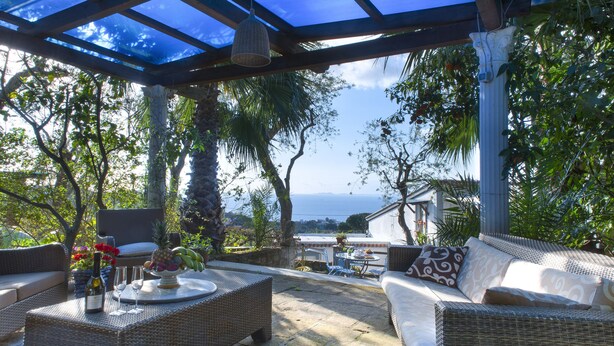 Un patio techado al aire libre con mobiliario de mimbre, una mesa con una botella y vasos, y vista al mar.