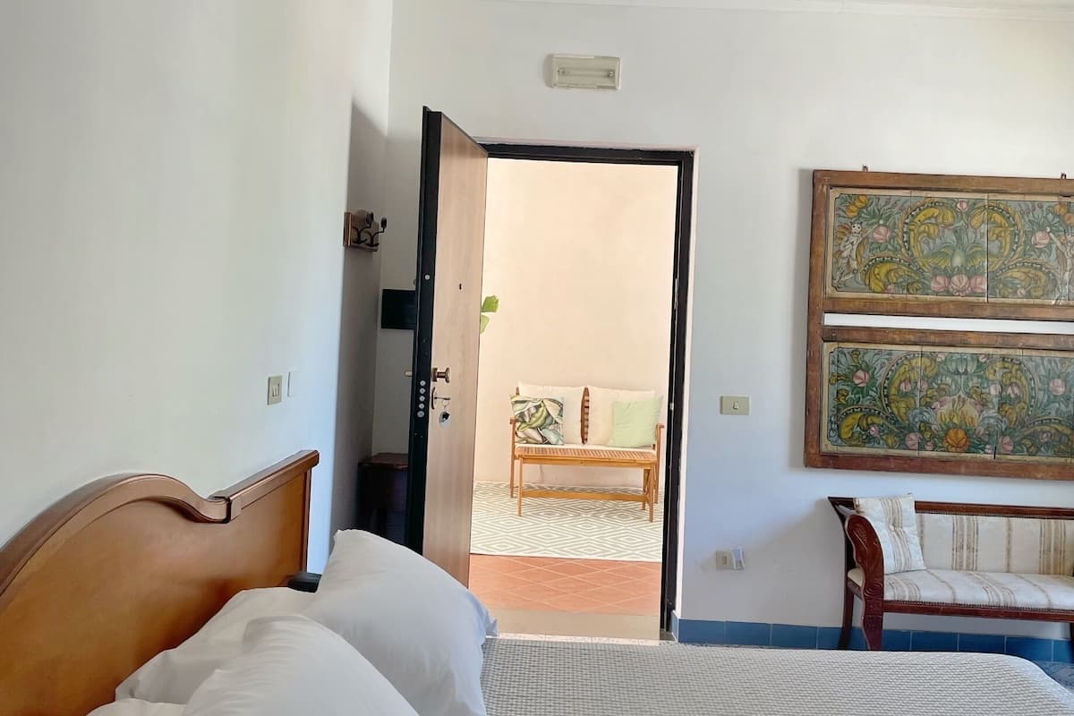 A hotel room with a large bed, a wooden bench, a wall-mounted painting, and a door leading to a balcony.