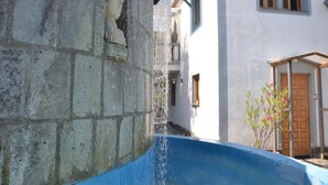 Un muro de piedra con una escultura en busto, una piscina azul y un edificio con puerta y ventanas de madera.