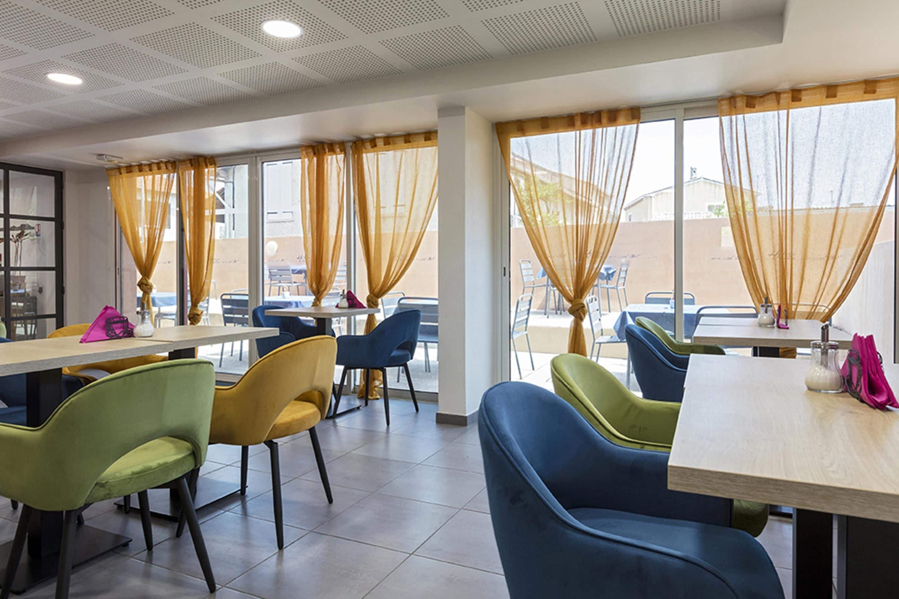 A dining area with tables and chairs, large windows with curtains, and a view of the outside.