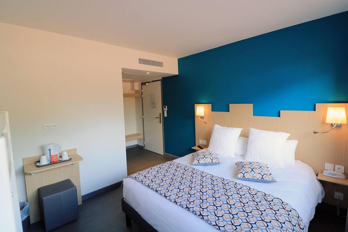 Habitación de hotel con cama, mesitas de noche, un escritorio pequeño y un cubo de basura.