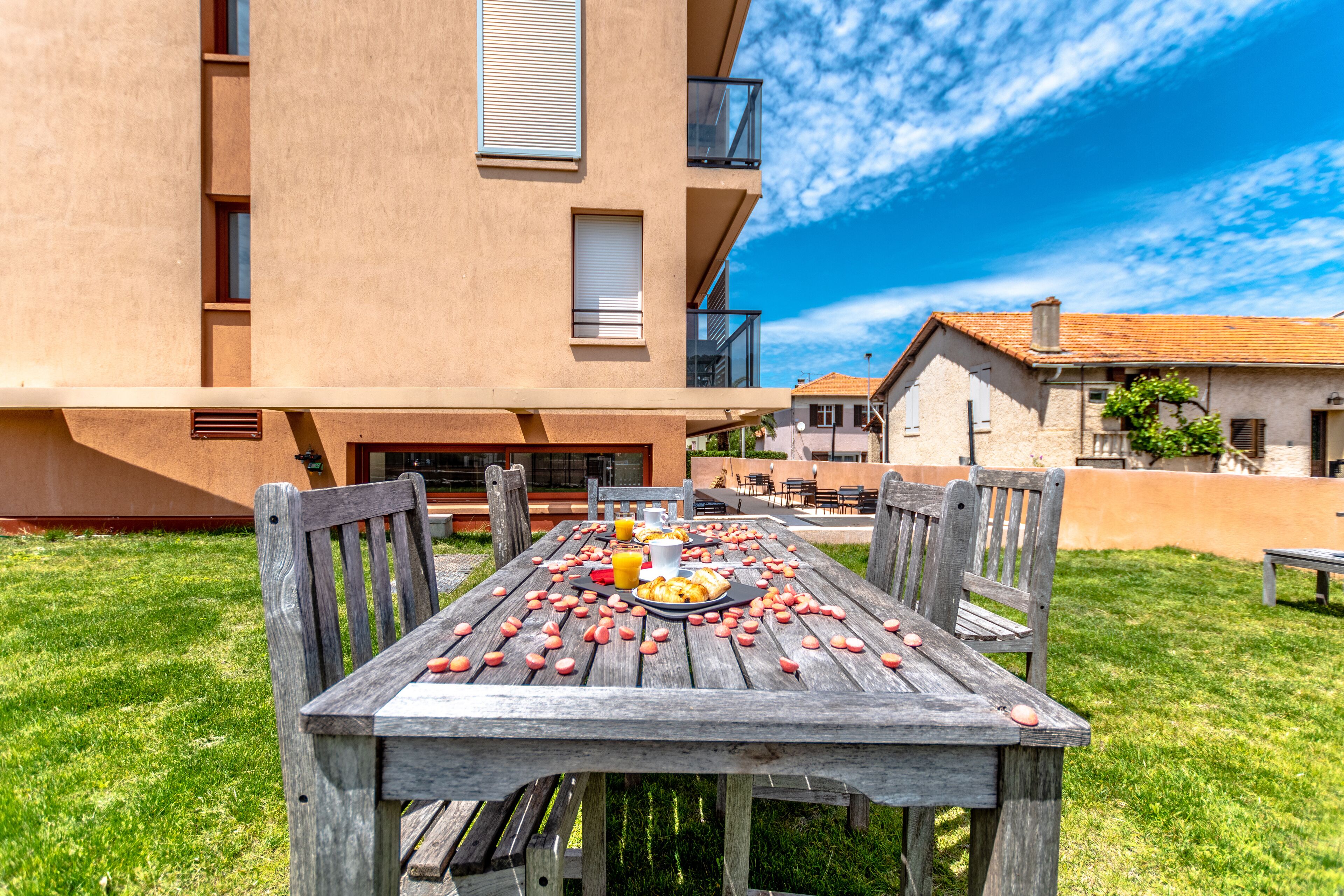 A wooden table set outdoors with food and drinks, surrounded by a grassy lawn and a building with balconies.