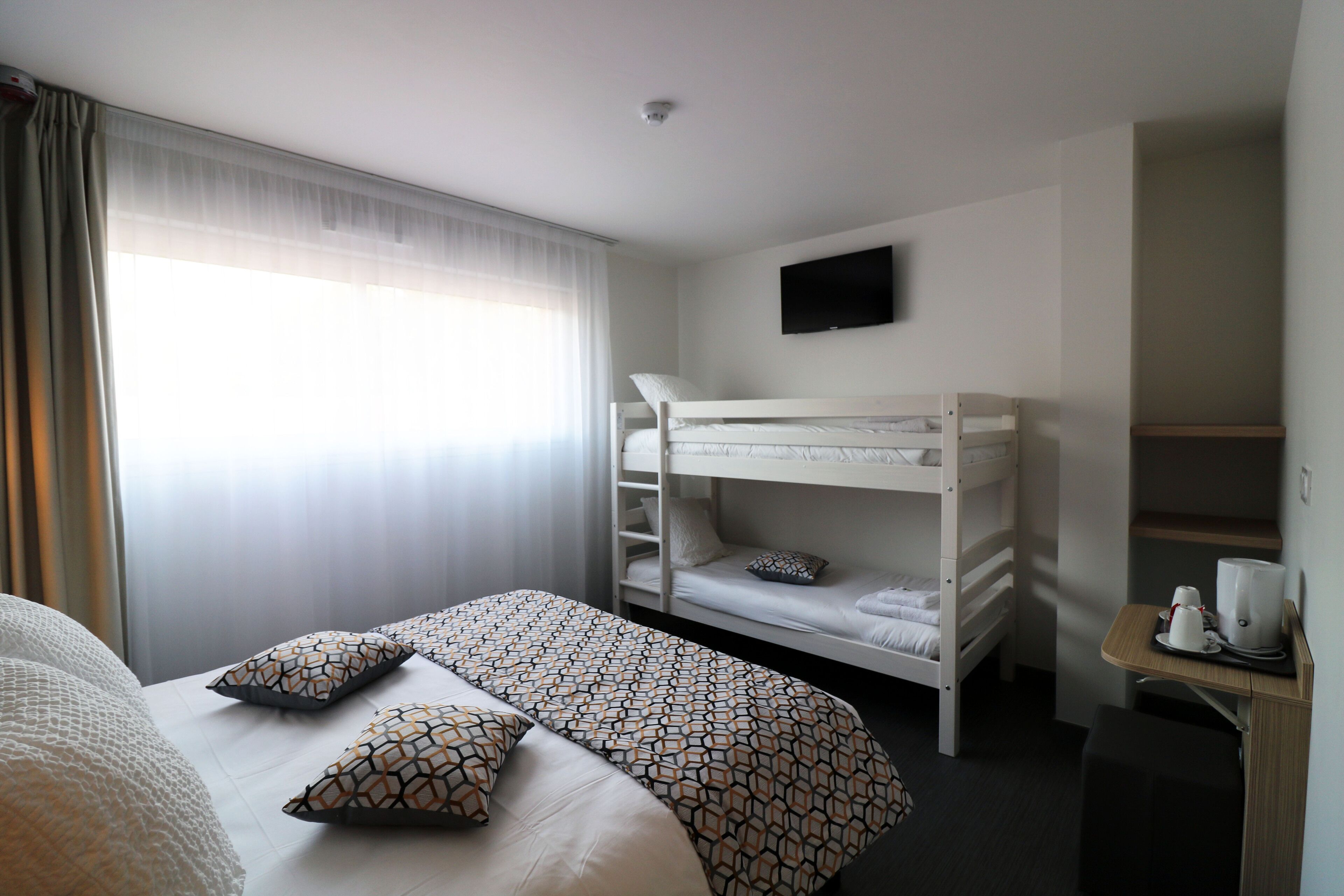 Une chambre d’hôtel avec un lit superposé, un lit simple agrémenté d’oreillers à motifs, un téléviseur fixé au mur et une petite table avec une machine à café.