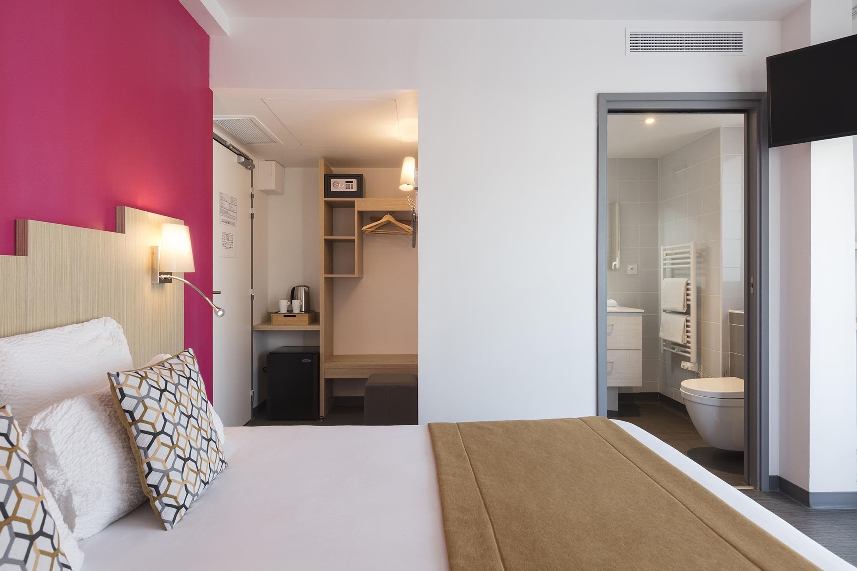 Een moderne hotelkamer met een bed, nachtkastje, garderobe en een badkamer zichtbaar door een geopende deur.
