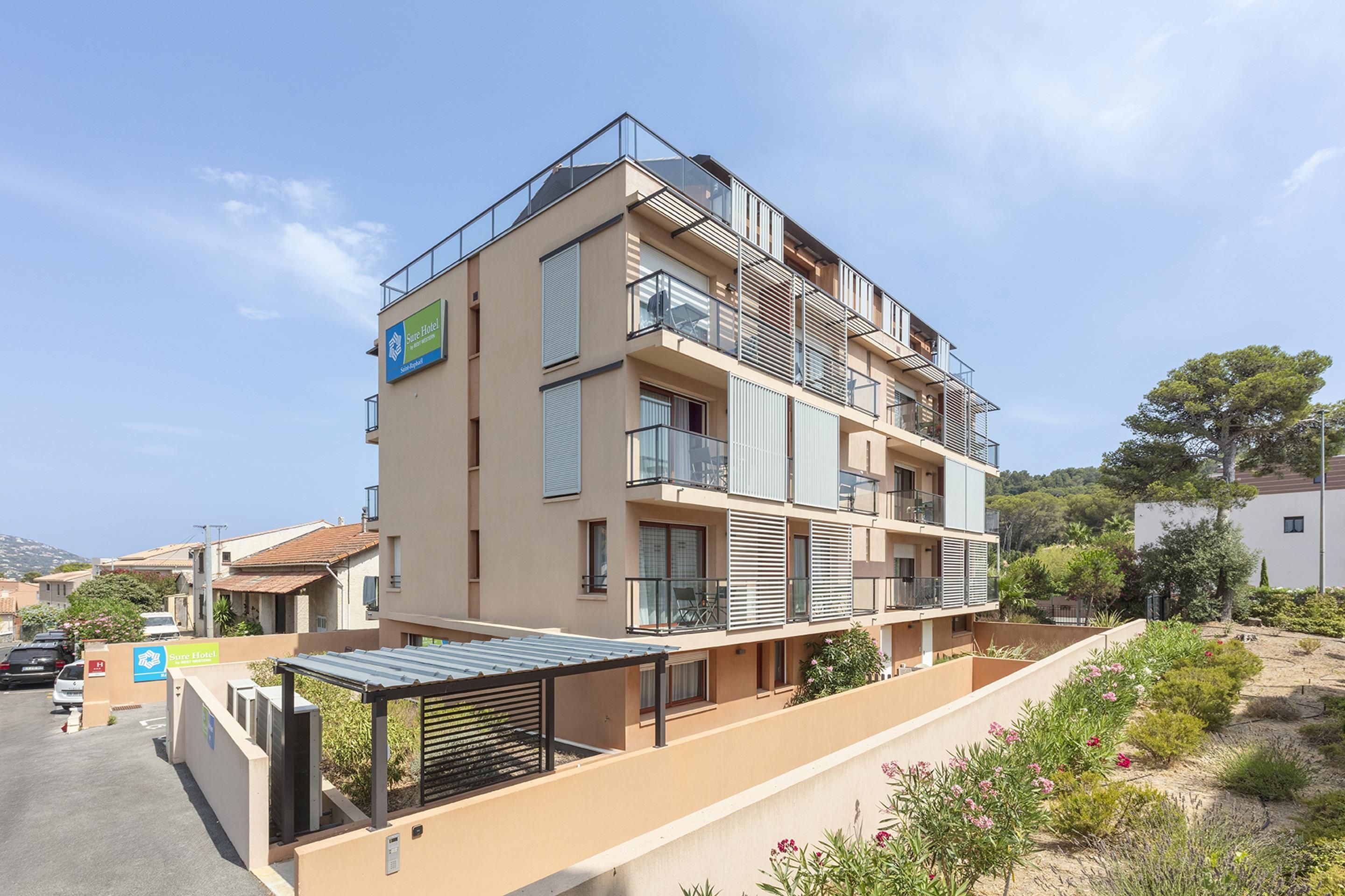 Un hôtel à plusieurs étages doté de balcons, d’un espace extérieur couvert aménagé en coin salon et d’un parking.