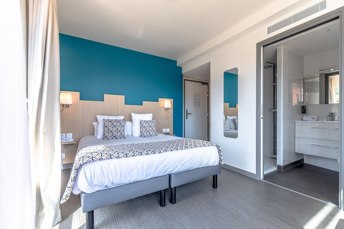 Habitación de hotel moderna con una cama grande, lámparas de noche, un baño con espejo y una puerta que da a otra habitación.