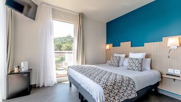A hotel room with a bed, a TV, a desk, a lamp, and a view of greenery outside.