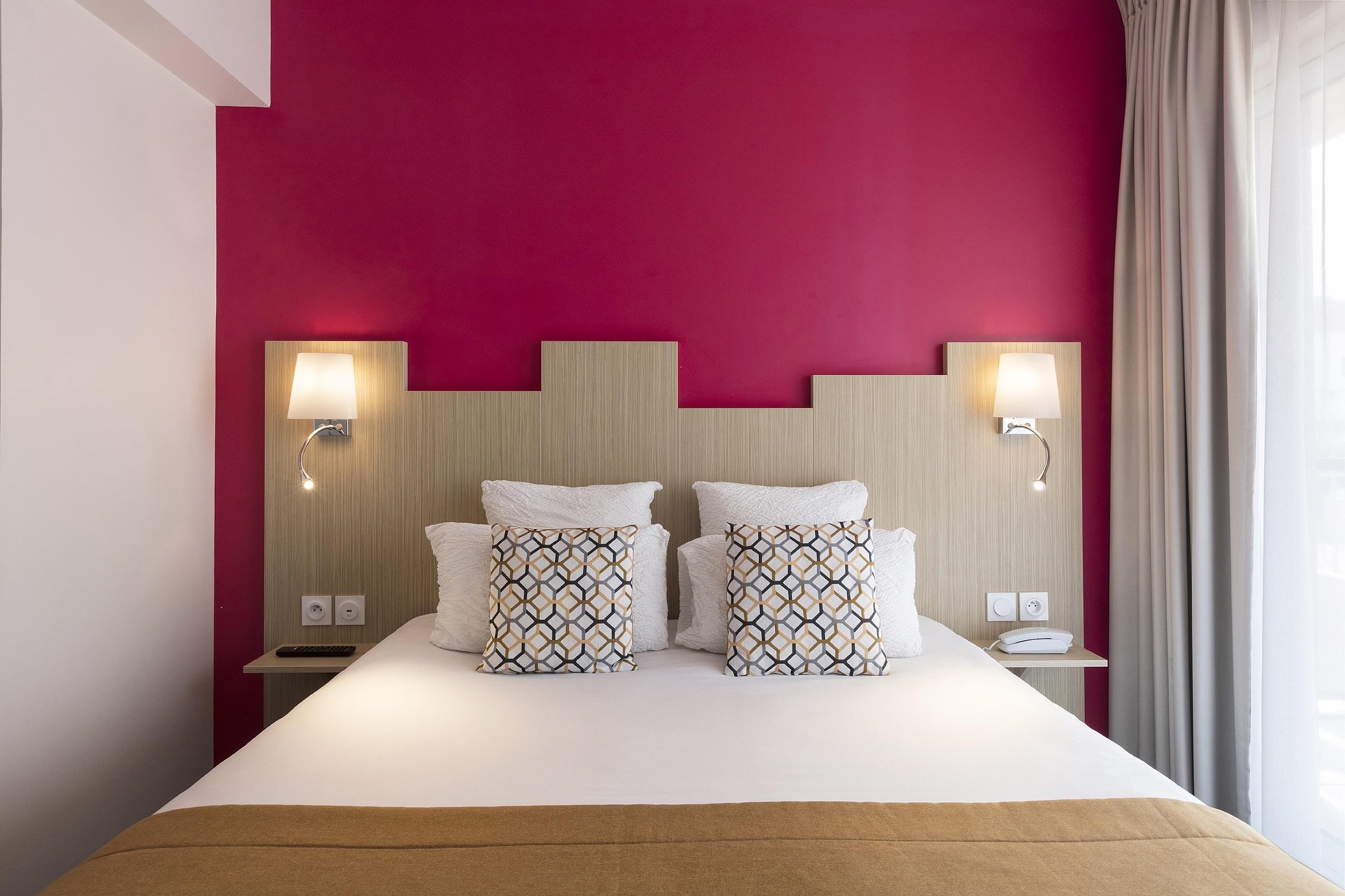 Habitación de hotel con cama, dos lámparas de noche, cabecera y ventana con cortinas.