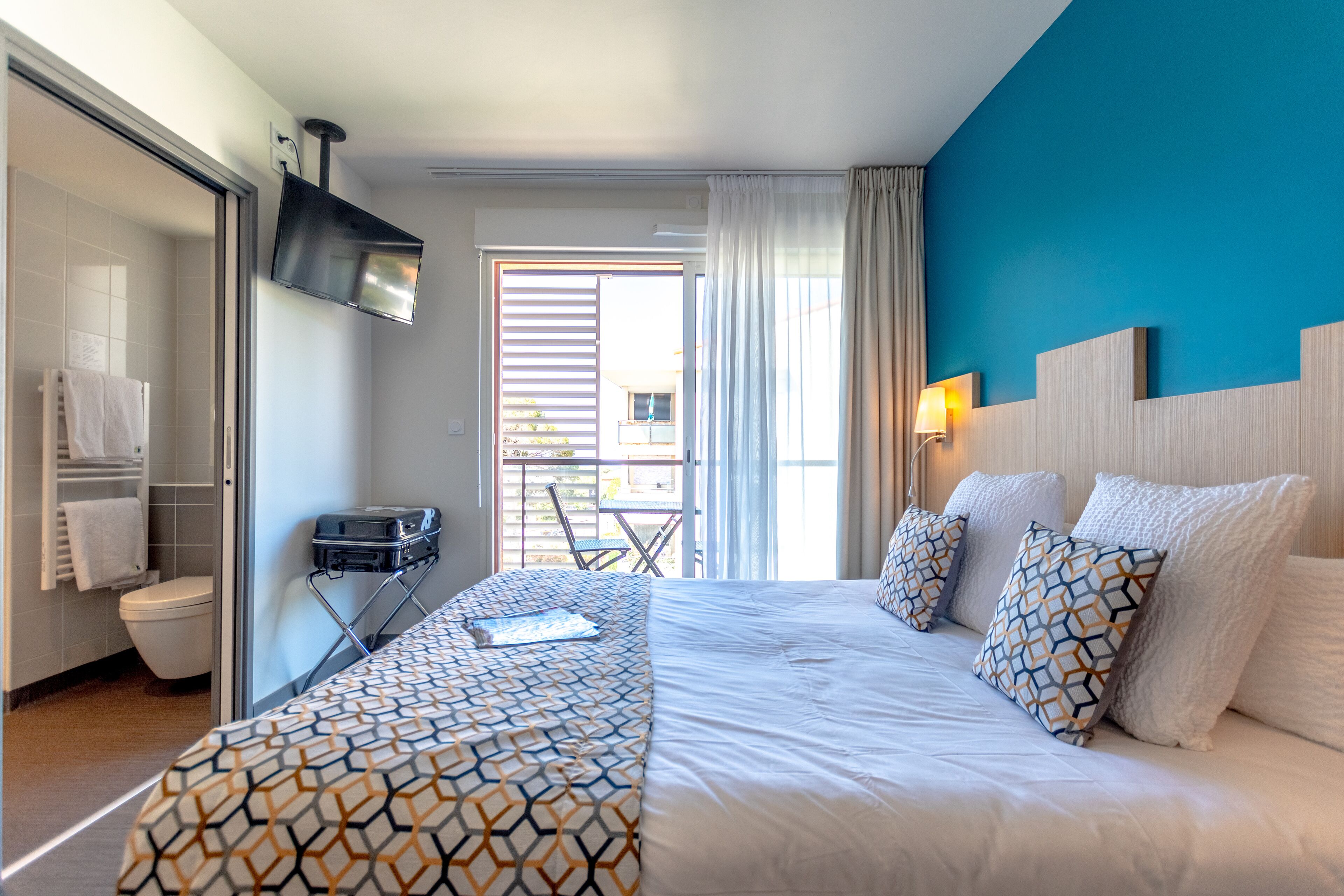 Une chambre d’hôtel comprenant un lit, un téléviseur fixé au mur, une salle de bain avec des toilettes et un porte-serviettes, et une vue sur l’extérieur par une fenêtre.