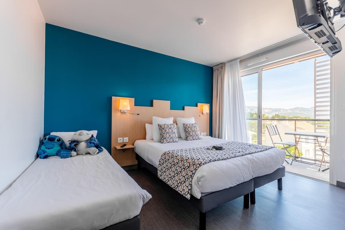 Habitación de hotel con una cama grande, otra más pequeña, un balcón con mesa y sillas, y vista al paisaje.