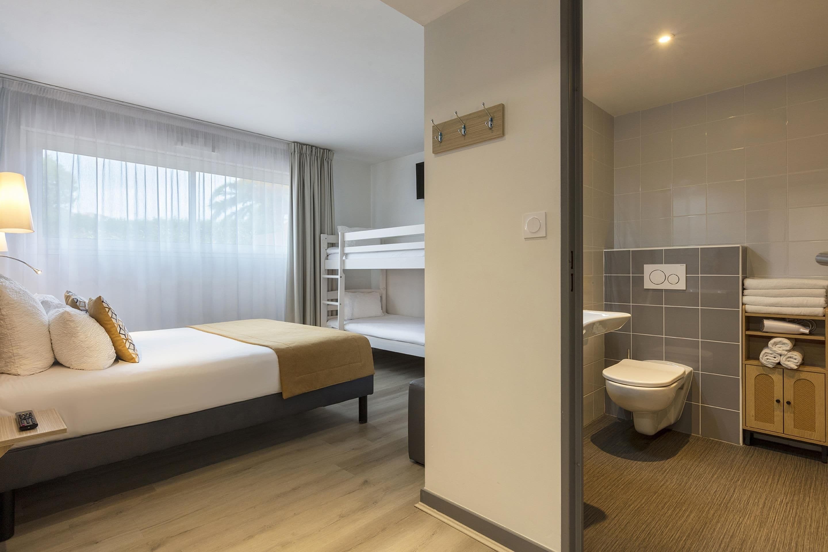 Een hotelkamer met een bed, nachtkastje, lamp, raam met gordijnen, een stapelbed, een badkamer met toilet, wastafel en handdoekrek.