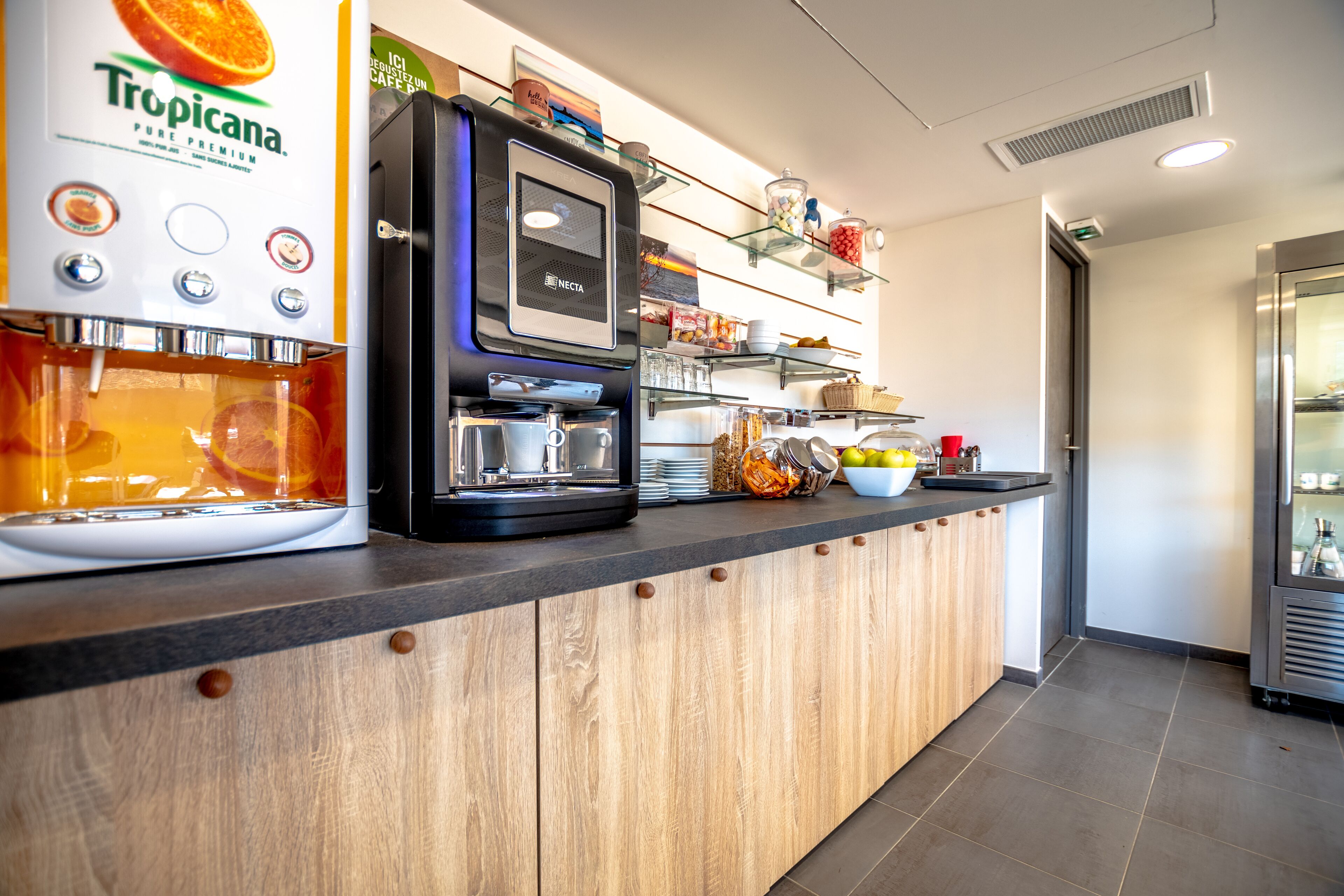 A self-service coffee station with a Tropicana juice dispenser, a Necta coffee machine, and various food items on shelves.