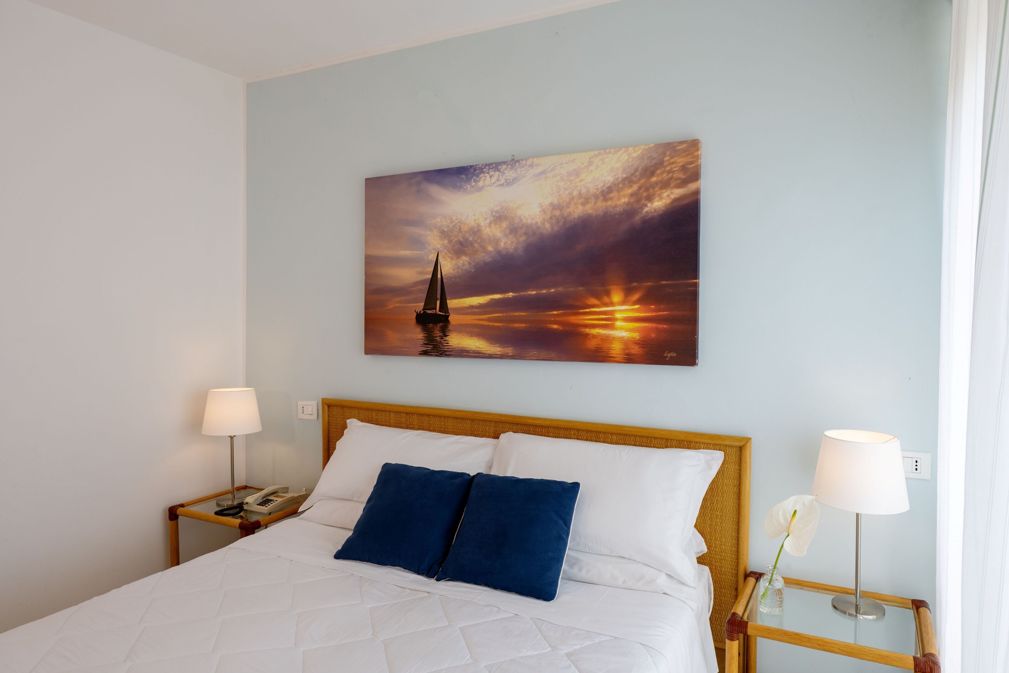 A hotel room with a bed, two bedside tables, a painting of a sunset, and a window with curtains.