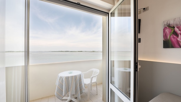 A balcony with a glass door, a small table with a striped tablecloth, and a white chair. - Capri (Grado)