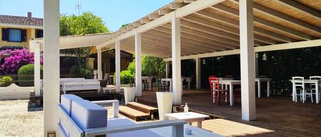Une terrasse extérieure avec des tables et des chaises en bois, un espace salon couvert et une vue sur un bâtiment en arrière-plan.