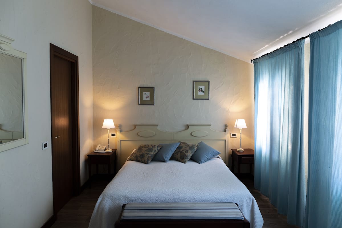 Une chambre d’hôtel avec un grand lit, deux lampes de chevet, une tête de lit agrémentée d’un miroir et une fenêtre avec des rideaux.