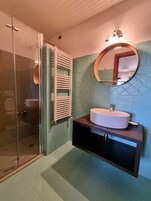 Un baño con cabina de ducha de vidrio, un lavabo blanco con mueble de madera y un espejo con marco redondo.