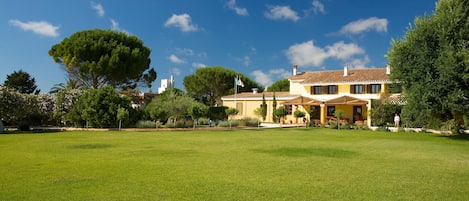 A large, well-maintained lawn in front of a yellow building with a tiled roof, surrounded by trees and a clear blue sky.