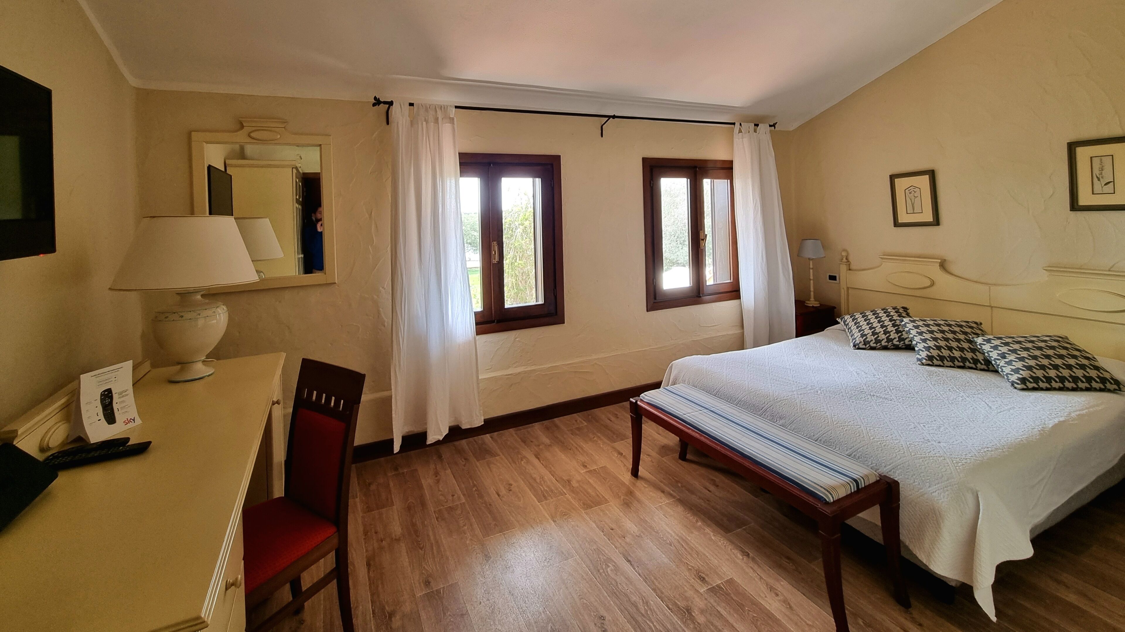 A hotel room with a bed, a desk with a lamp, a chair, and two windows with curtains.