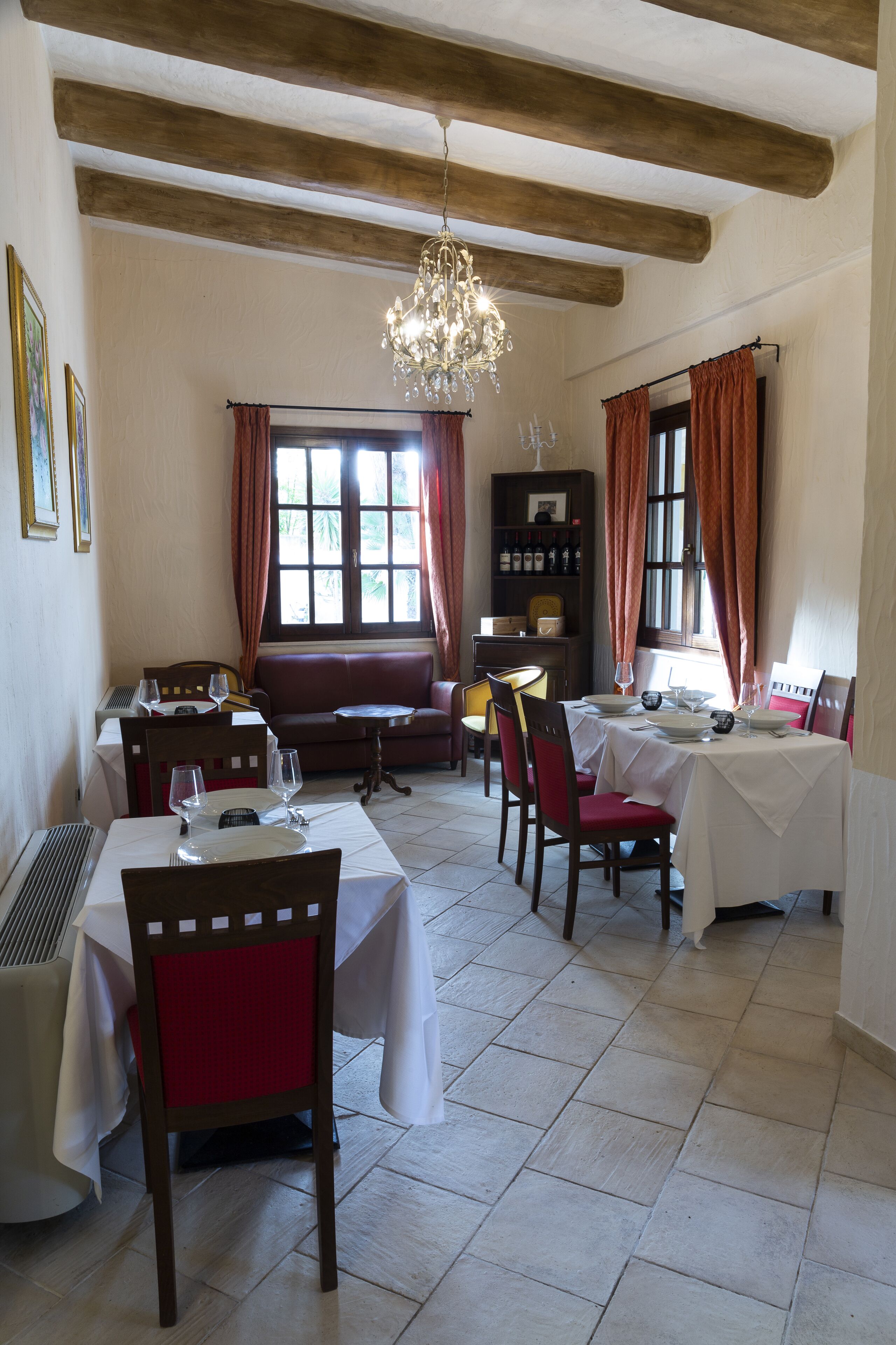 A dining area with tables set for four, a chandelier, and a bookshelf with wine bottles.