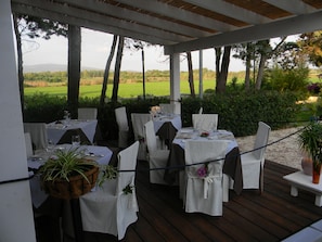 Un área de comedor al aire libre con mesas dispuestas para dos, rodeada de sillas y cubierta con manteles blancos.