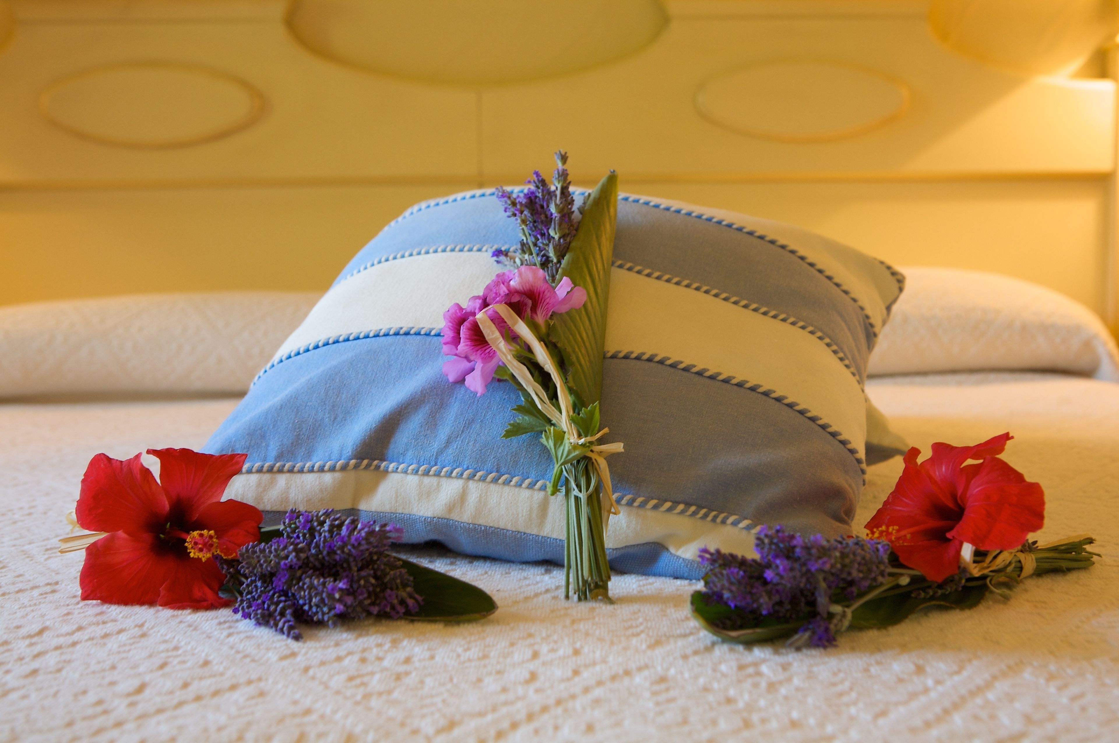 Una cama bien hecha con un cojín azul y blanco a rayas, un ramo de flores y dos flores de hibisco rojo sobre la colcha.