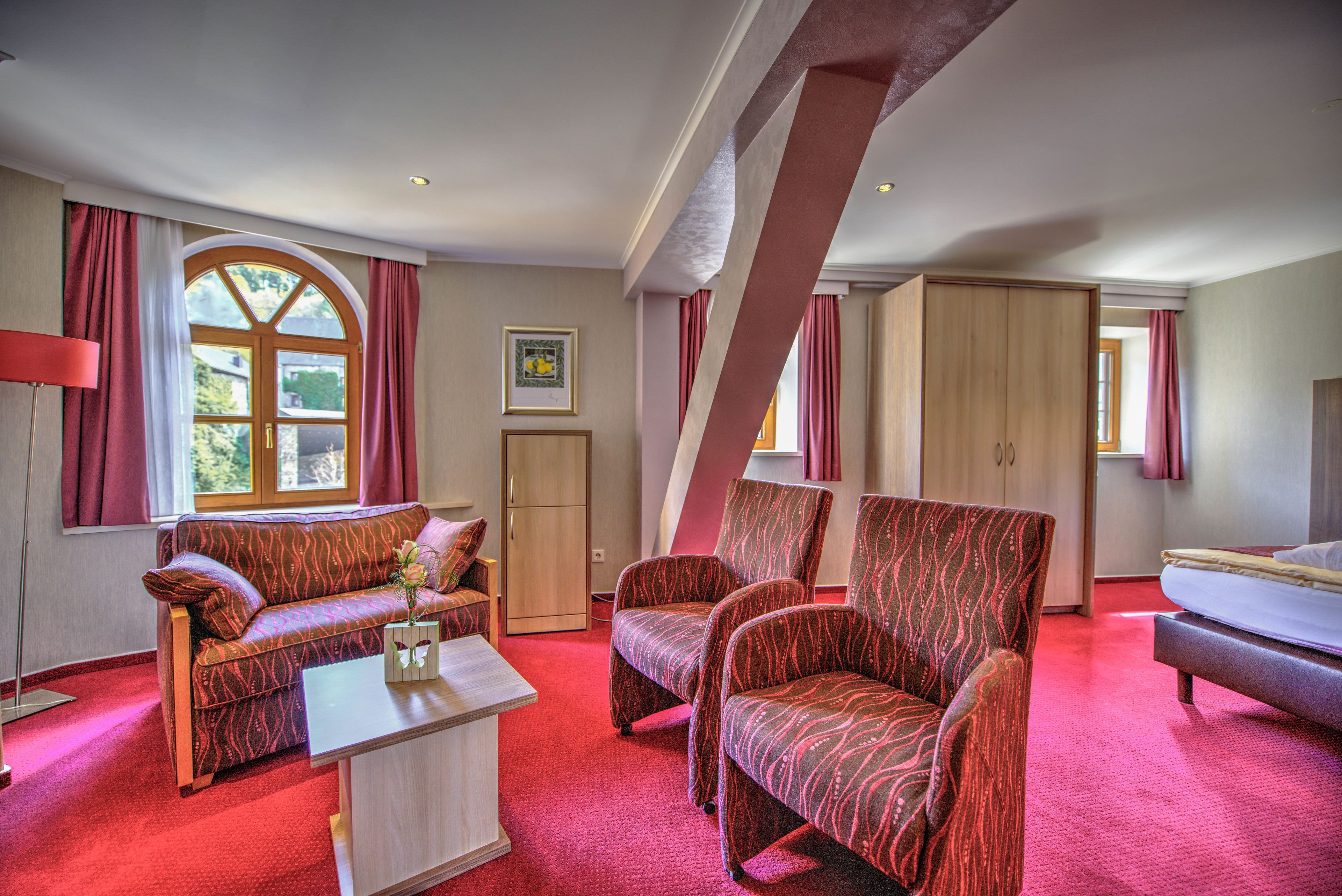 Habitación de hotel con alfombra roja, un sofá, dos sillones, una cama, un armario de madera y un ventanal con cortinas.