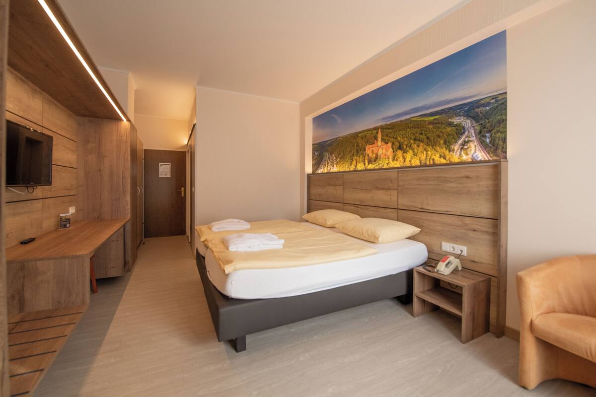 A modern bedroom with a large bed, a wooden headboard, a nightstand, a chair, and a painting of a landscape on the wall.