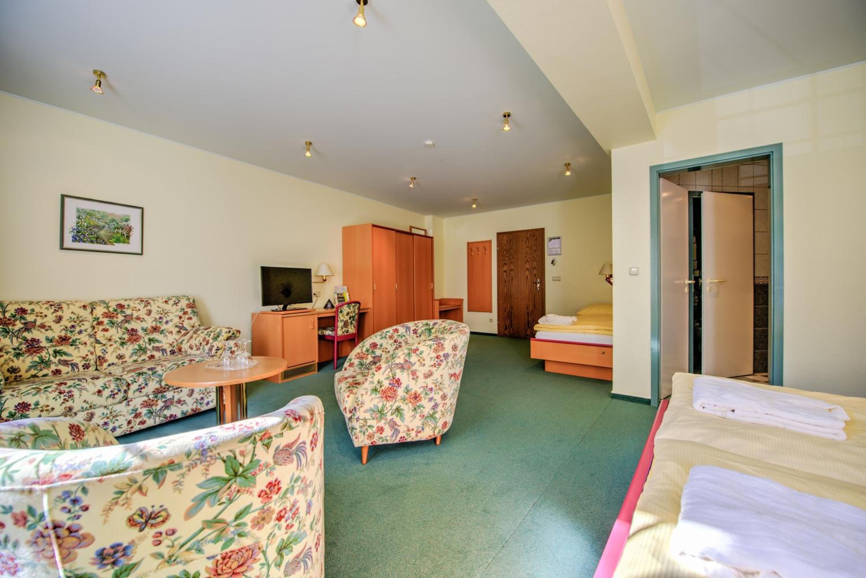 Habitación de hotel con un sofá floral, una pequeña mesa redonda, un armario de madera, una cama con cabecera rosa y una puerta que conduce a otra habitación.