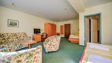 Habitación de hotel con un sofá floral, una pequeña mesa redonda, un armario de madera, una cama con cabecera rosa y una puerta que conduce a otra habitación.