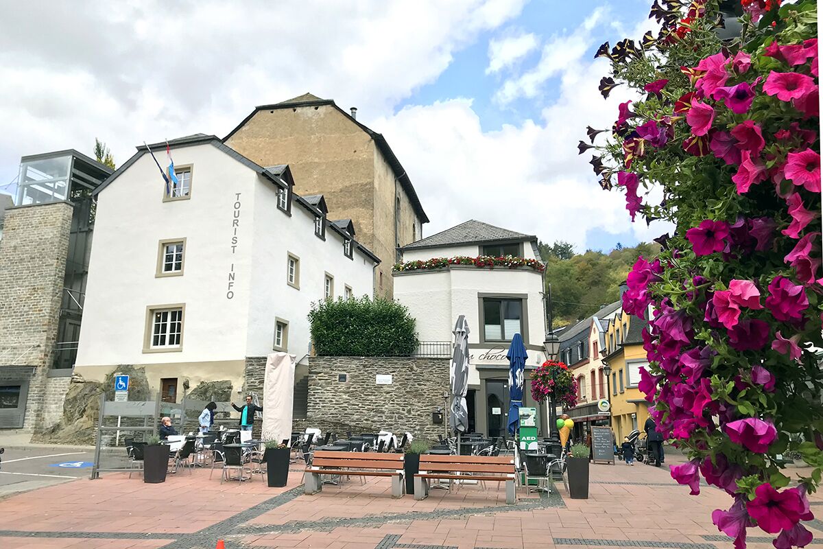 Ein weißes Gebäude mit einem Schild, auf dem 'TOURIST INFO' steht, und eine Steinmauer. Es gibt Tische und Bänke im Freien sowie eine Blumenpracht.