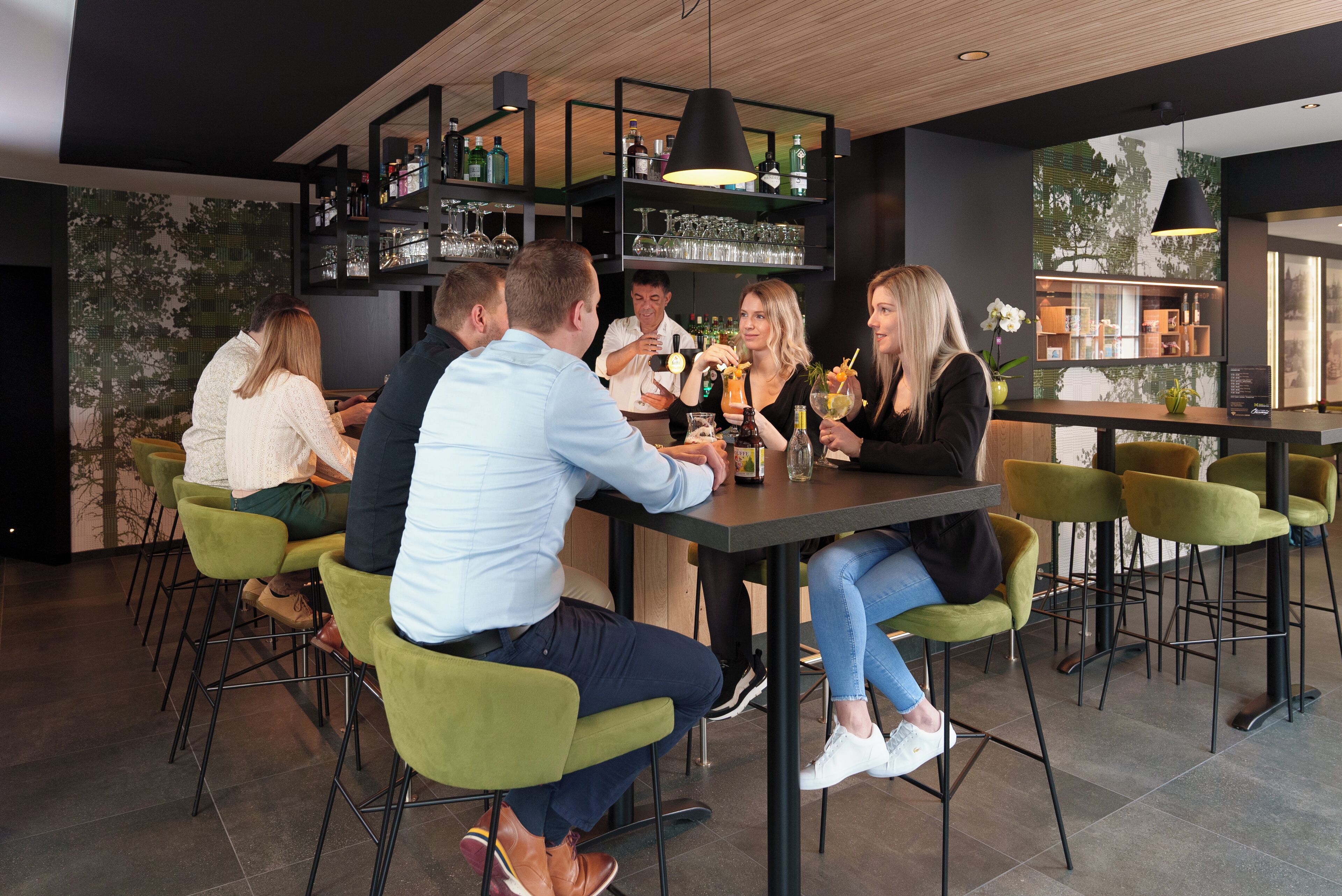 Un área de bar moderna con un grupo de personas sentadas alrededor de una mesa, una barra con estantes llenos de botellas y una pared verde con un mural de un árbol.