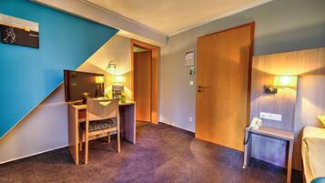 A hotel room with a desk, chair, and a small table with a lamp. There is a door and a wall with a blue accent.
