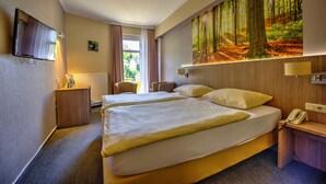 A hotel room with a large bed, a desk with a telephone, a TV mounted on the wall, and a painting of a forest.