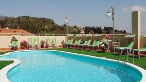 Piscina exterior con césped verde, rodeada de sillas de descanso y vistas a una colina arbolada.