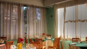 Comedor con mesas dispuestas para cuatro, sillas de madera y vistas a través de cortinas translúcidas.