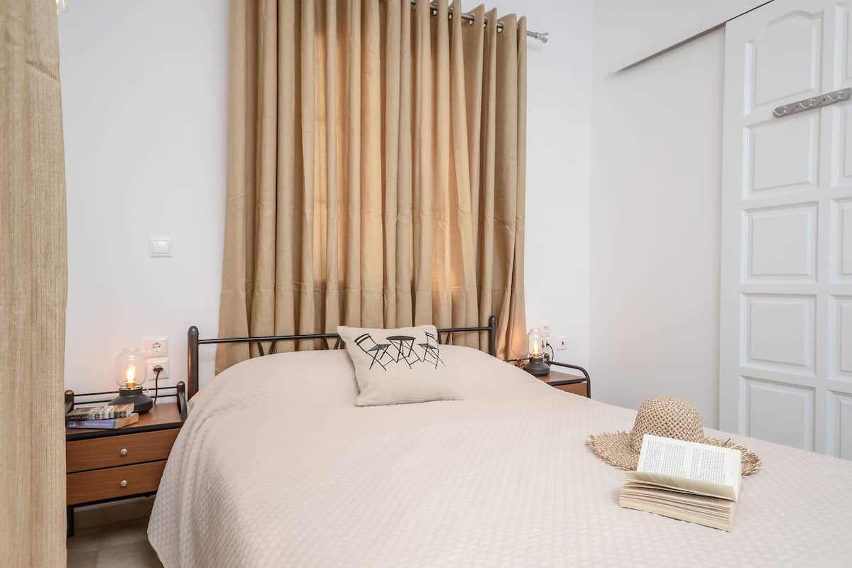 A neatly made bed with a beige bedspread, a pillow with a chair design, a bedside lamp, and a book on the nightstand.