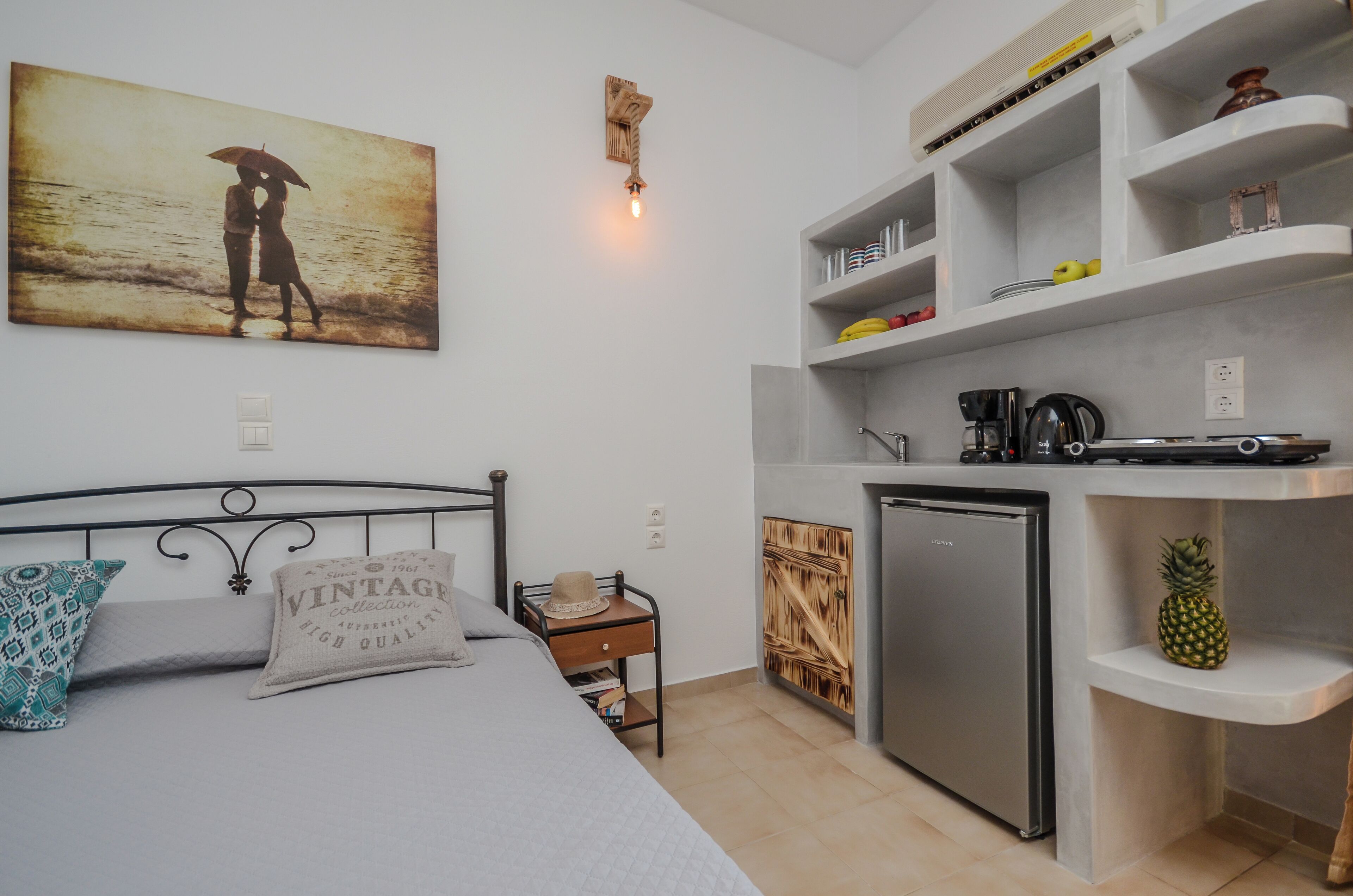 Quarto compacto com cama, uma pequena mesa de cabeceira, cozinha compacta equipada com geladeira e prateleira fixada na parede com diversos objetos.