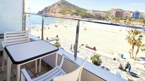 Un balcón con mesa y sillas, con vista a una playa donde se ven personas y edificios al fondo.