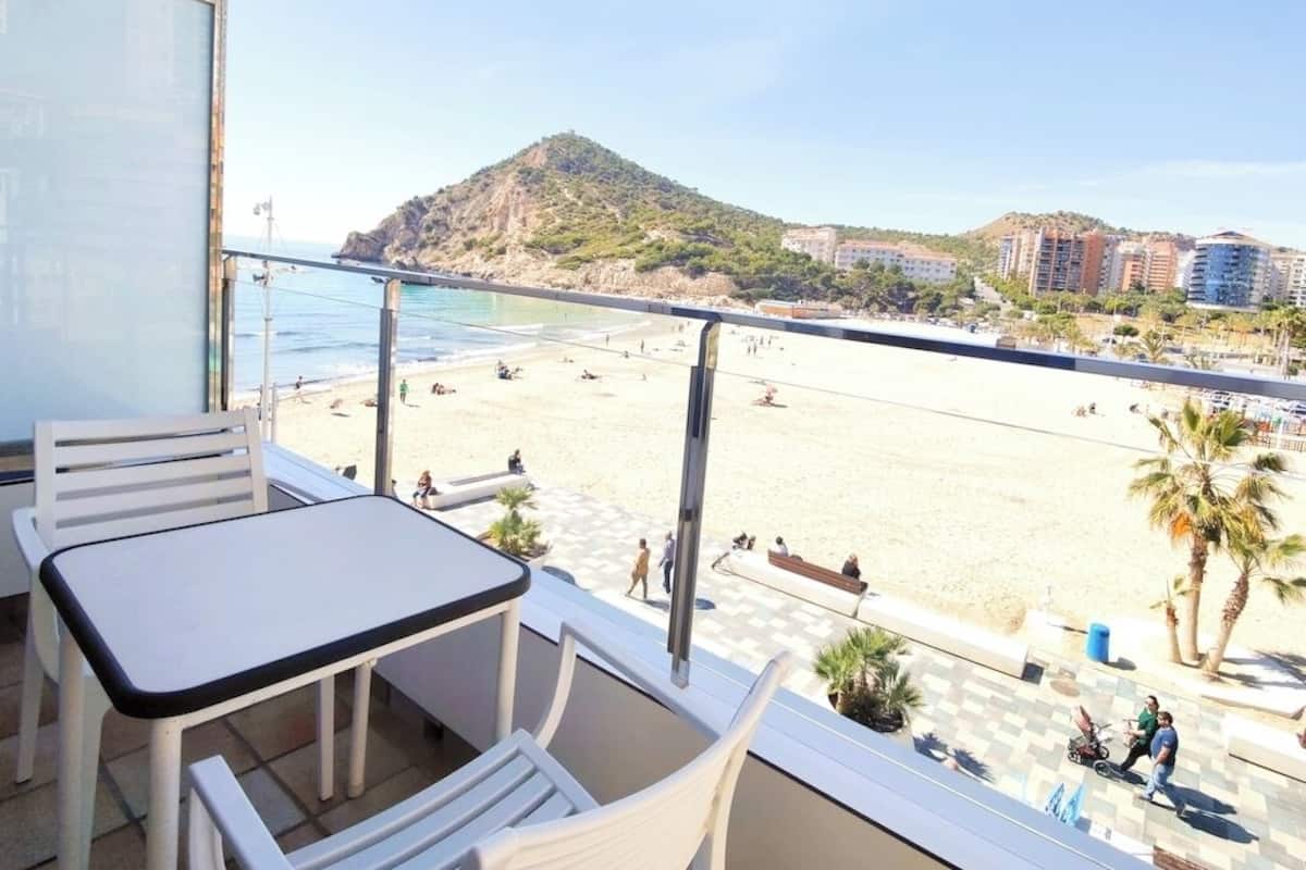 A balcony with a table and chairs, overlooking a beach with people and buildings in the background.