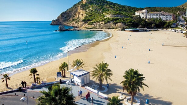 Una playa con vista despejada al mar, un pequeño quiosco, palmeras y un edificio al fondo.