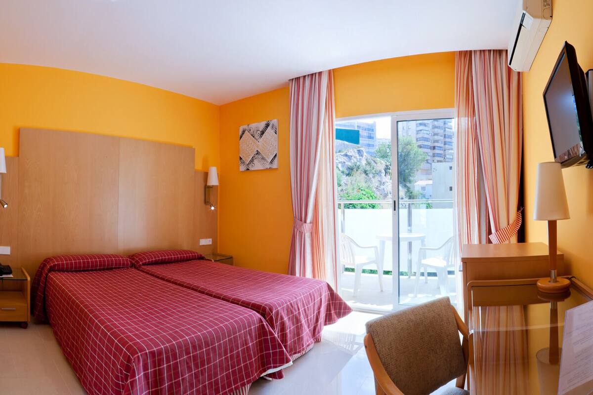 A hotel room with two beds, a desk, a chair, a TV, and a balcony with a view of buildings and greenery.