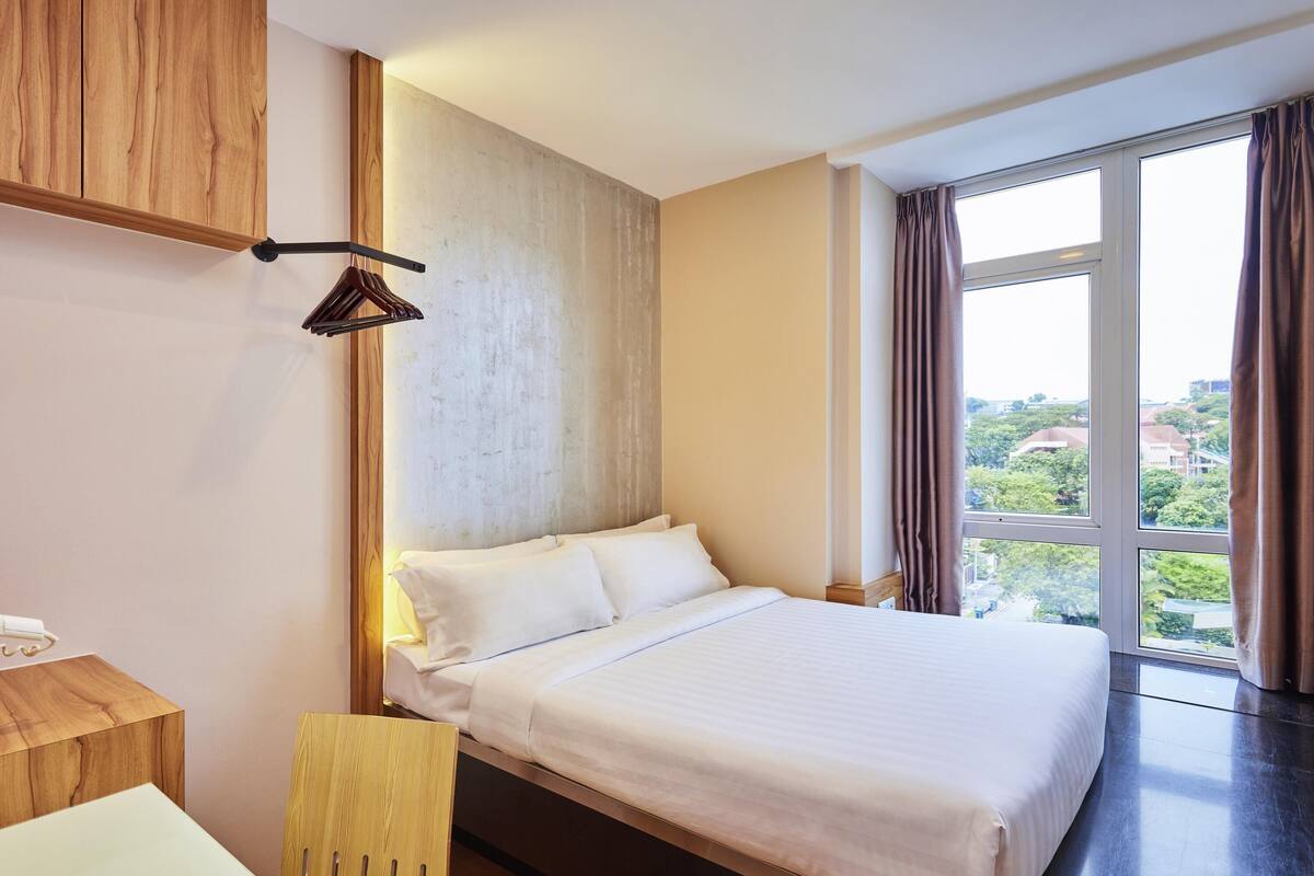 A modern hotel room with a large bed, a wooden desk, and a view of the city through large windows.