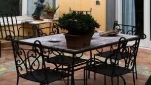 Un patio al aire libre con una mesa redonda y sillas, rodeado de plantas en macetas y una lámpara de pared decorativa.