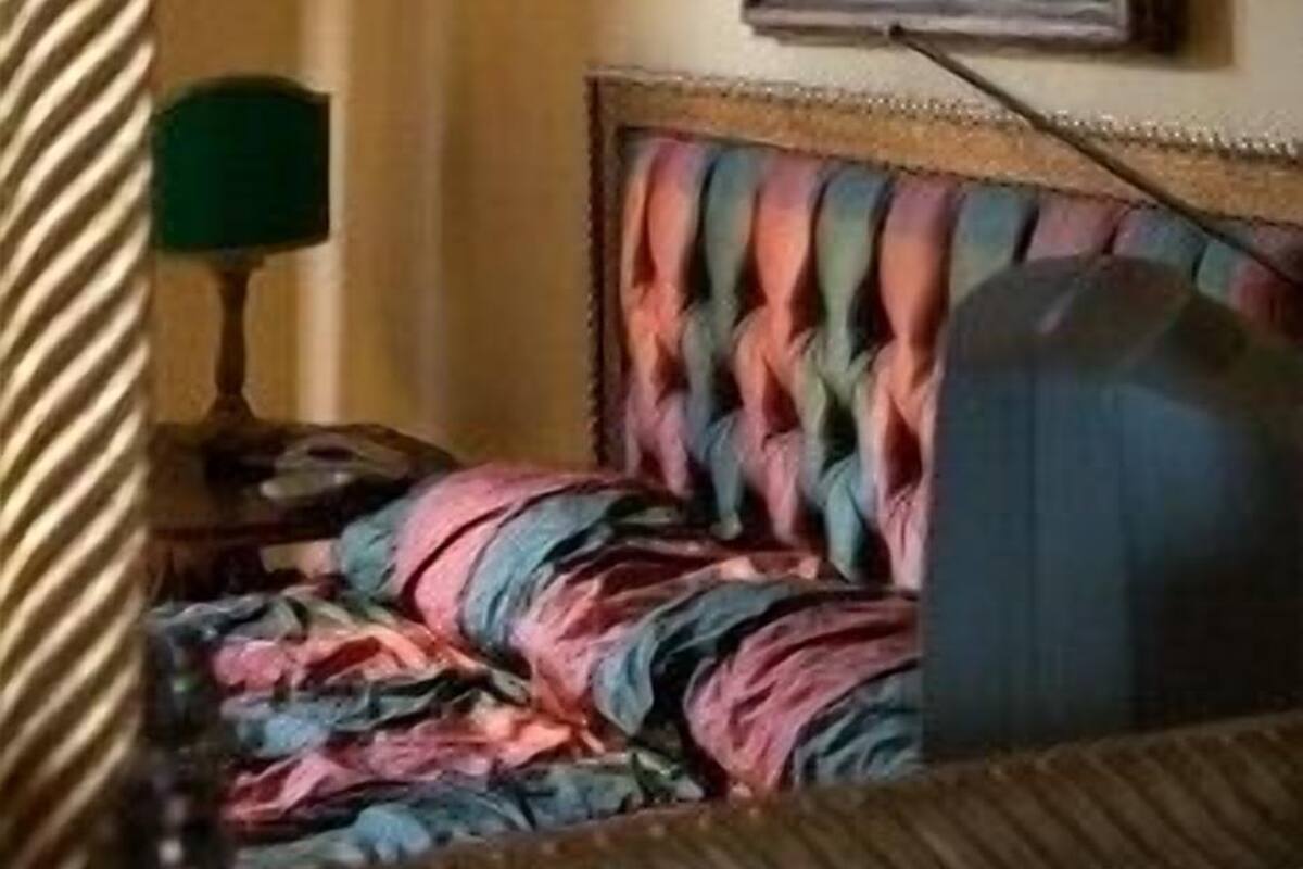 A hotel room with a bed featuring a colorful, patterned headboard, a painting on the wall, and a green lamp on a bedside table.