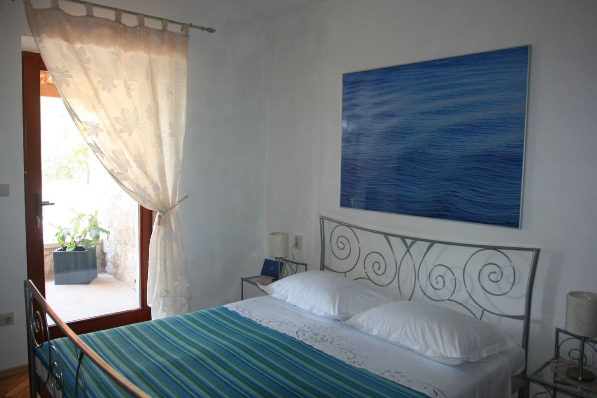 A bedroom with a bed, a window with curtains, a bedside table with a lamp, and a painting on the wall.