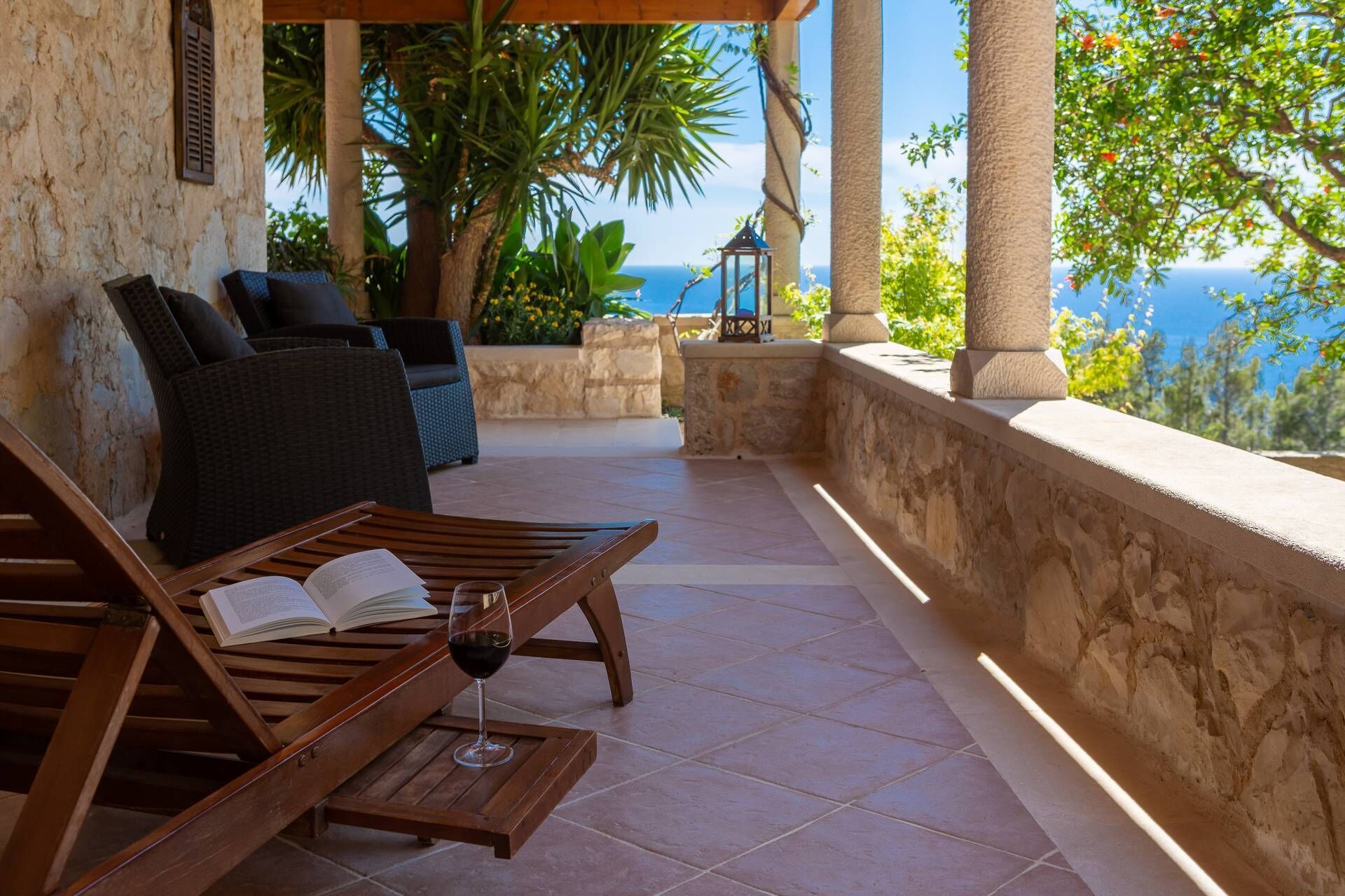 A wooden lounge chair with a book and a glass of red wine, a wicker sofa, and a view of the sea.